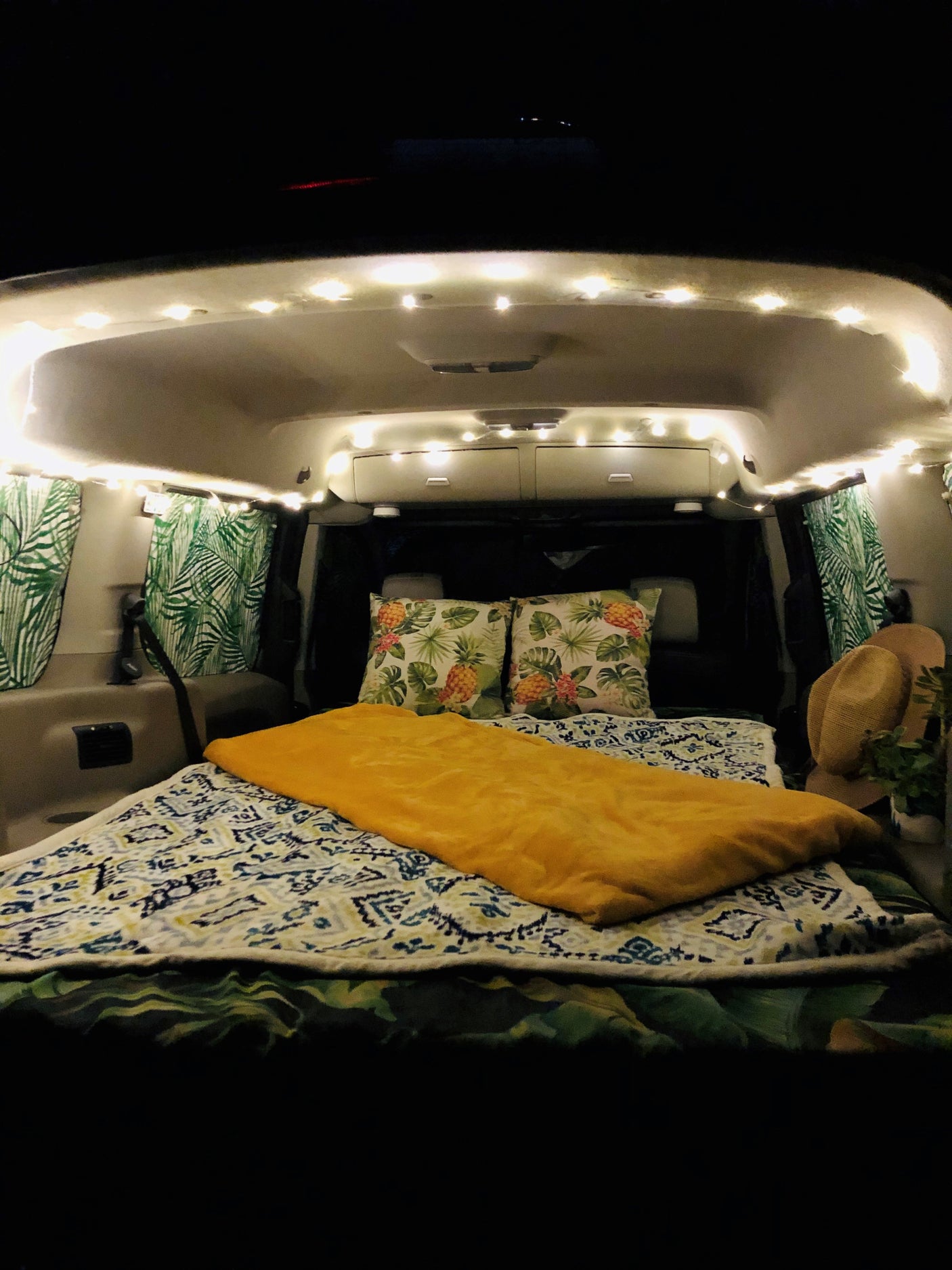 Cozy RV rental interior at night. A comfy bed with patterned blankets, pineapple pillows, and palm-leaf curtains are illuminated by string lights.