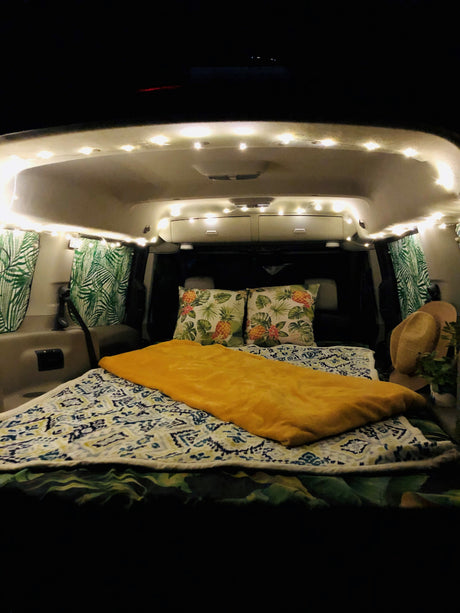 Cozy RV rental interior at night. A comfy bed with patterned blankets, pineapple pillows, and palm-leaf curtains are illuminated by string lights.
