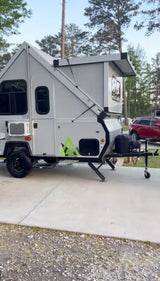 A light gray A-frame pop-up camper, a great rv rental, set up on concrete in a wooded campsite.