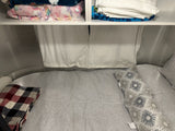 Cozy RV rental sleeping nook: a bed with light gray linens, a plaid blanket, pillows, and shelves with folded blankets/towels.