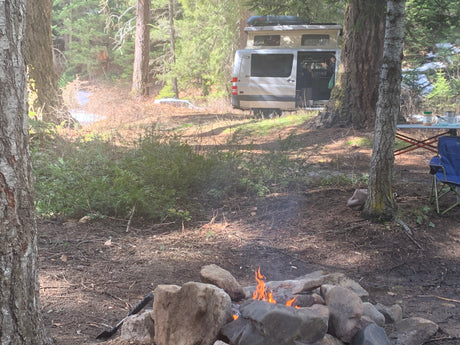 Enjoying a cozy RV rental campsite with a crackling campfire, parked campervan, and a person visible in the forest.