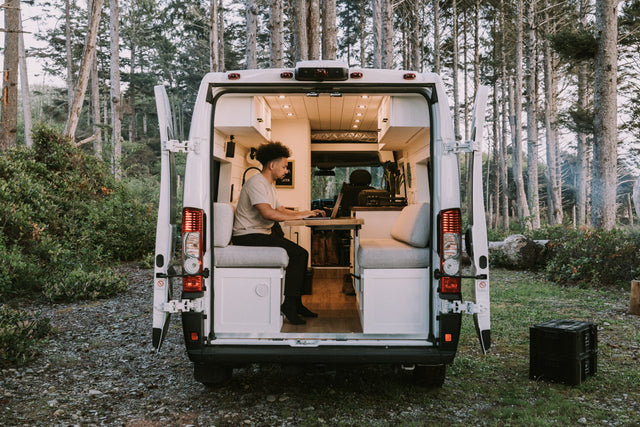 Man works on laptop in a modern RV rental camper van, parked in a serene forest. Ideal for remote work & travel.