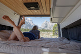 Enjoy an RV rental adventure! A person relaxes on a cozy bed inside a van, gazing at majestic mountain scenery.