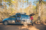 Exploring in an RV rental minivan. Parked on a dirt road, open doors reveal a custom camper setup; two people access the trunk.