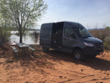 Gray Mercedes Sprinter RV rental van parked on red sand next to a lake and picnic table. A person is visible inside.