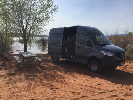 Gray Mercedes Sprinter RV rental van parked on red sand next to a lake and picnic table. A person is visible inside.