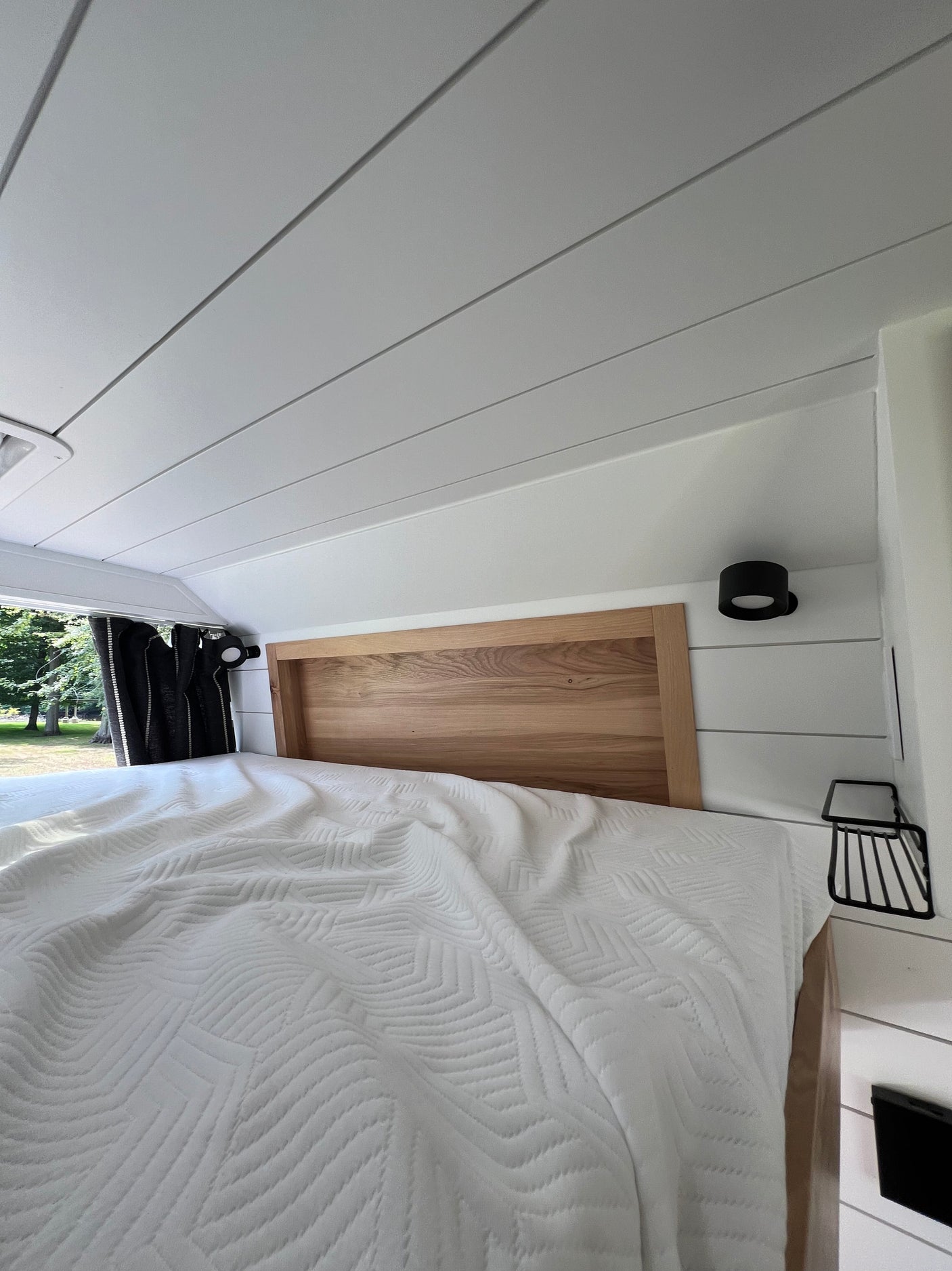 Cozy RV rental sleeping area: bed with white patterned comforter, wooden headboard, shiplap walls, and window view.