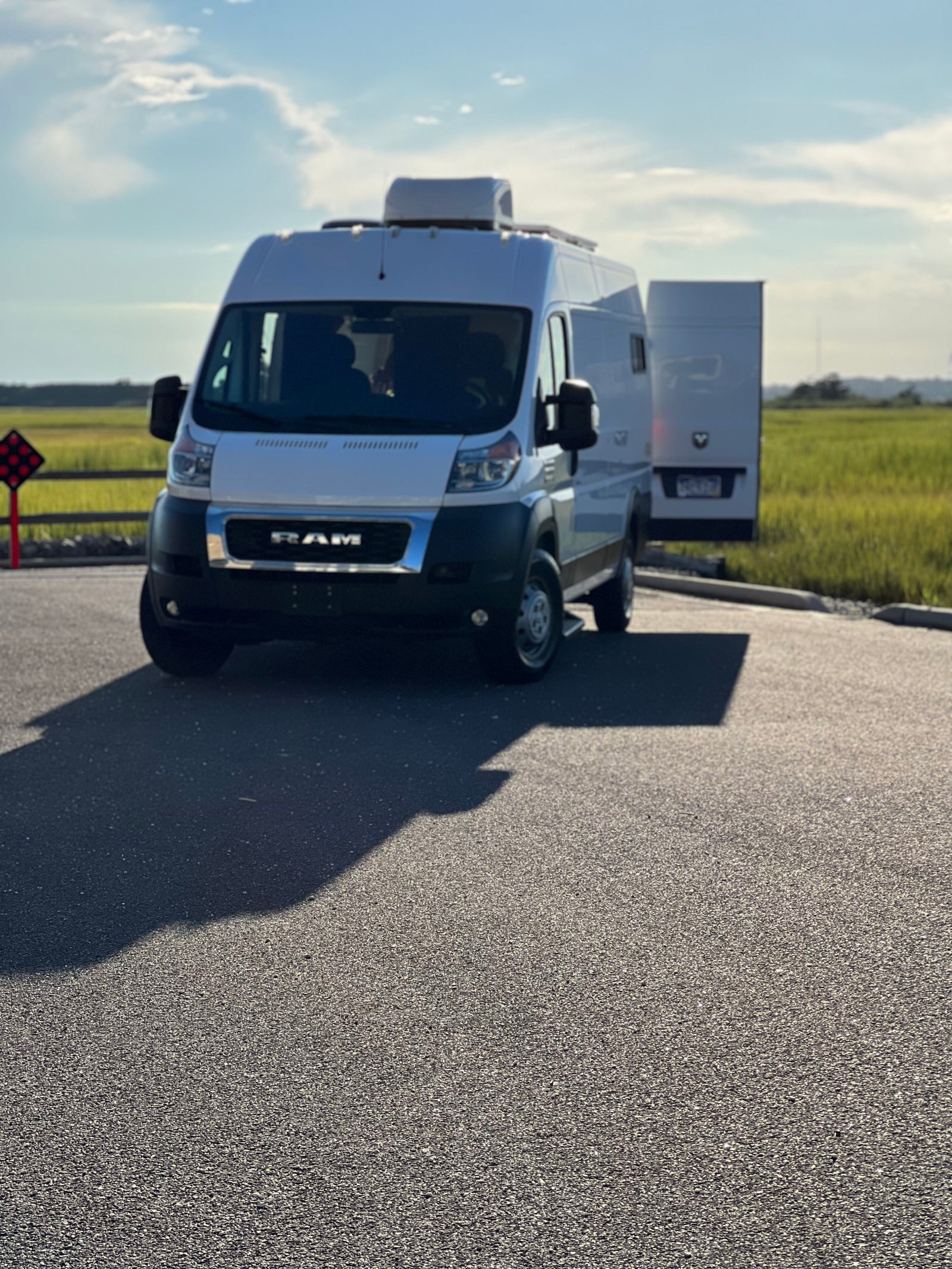 White Ram ProMaster RV rental van parked in a sunny lot, ready for adventure. Grassy field and blue sky in background.