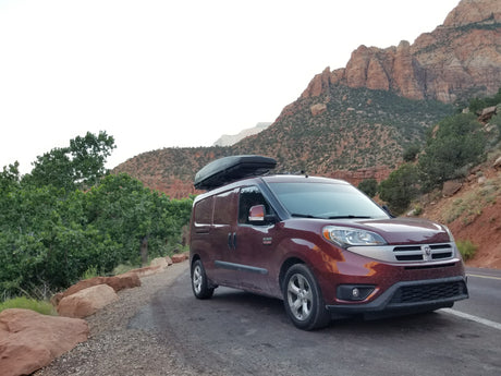 A burgundy Ram van, perfect for an RV rental adventure, parked by red rock mountains & green foliage.