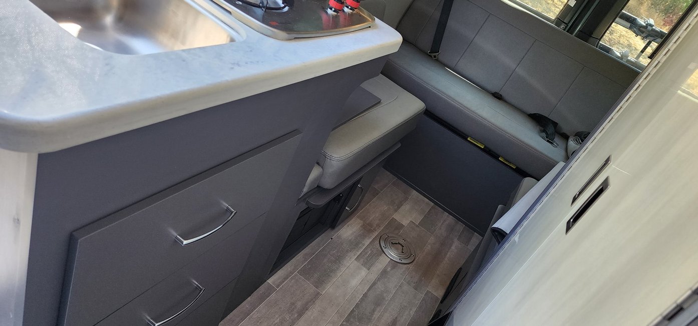 Functional RV rental interior with kitchen counter, sink, and dark grey cabinetry. Adjacent is a comfy grey bench seat, all on wood-look flooring.