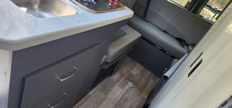 Functional RV rental interior with kitchen counter, sink, and dark grey cabinetry. Adjacent is a comfy grey bench seat, all on wood-look flooring.