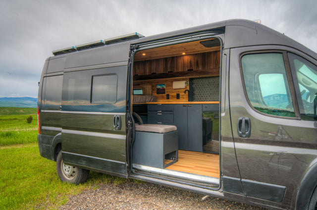 This RV rental offers a grey camper van with an open door revealing a modern interior, kitchen, & bed, in nature.