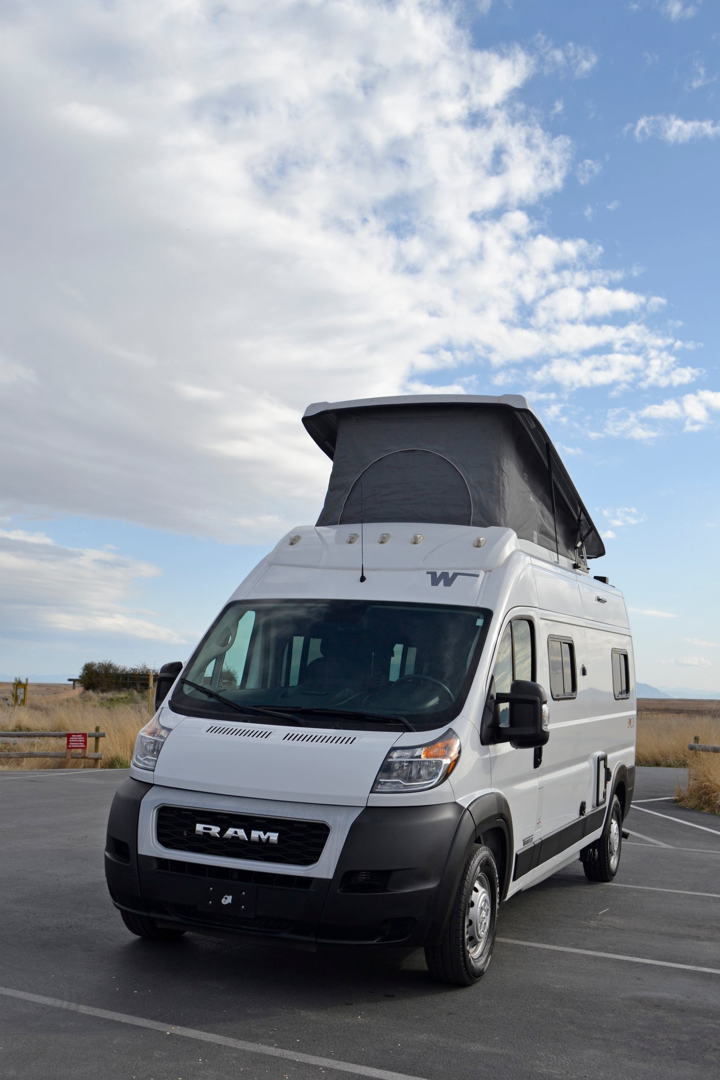 White camper van RV rental with pop-top extended, parked in a lot against a dry, grassy landscape and blue sky.