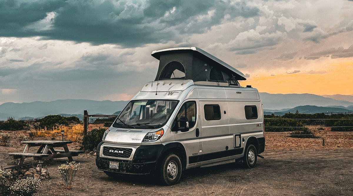 Silver camper van RV rental parked at a scenic mountain overlook. Dramatic sunset sky over mountains for a nature escape.|Silver pop-top RV rental