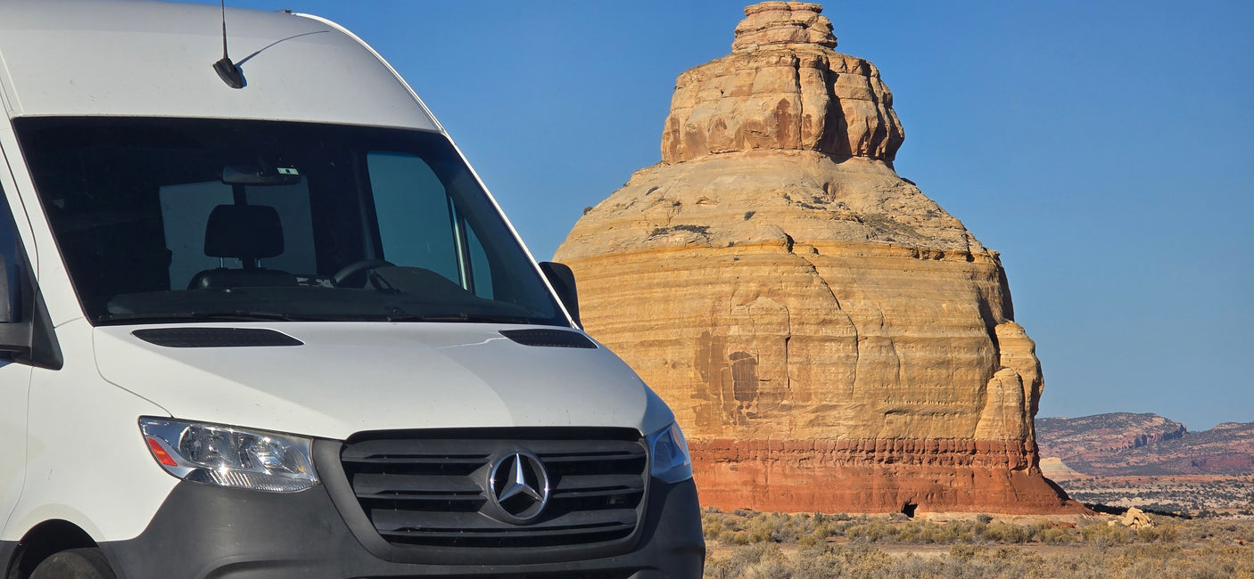 A white Mercedes Sprinter rv rental is parked in a desert landscape before a majestic, layered rock formation under a clear sky.