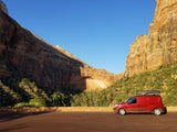 Red **rv rental** van with roof box, parked before towering, sunlit canyon walls. Perfect for **road trips** & **van life**.