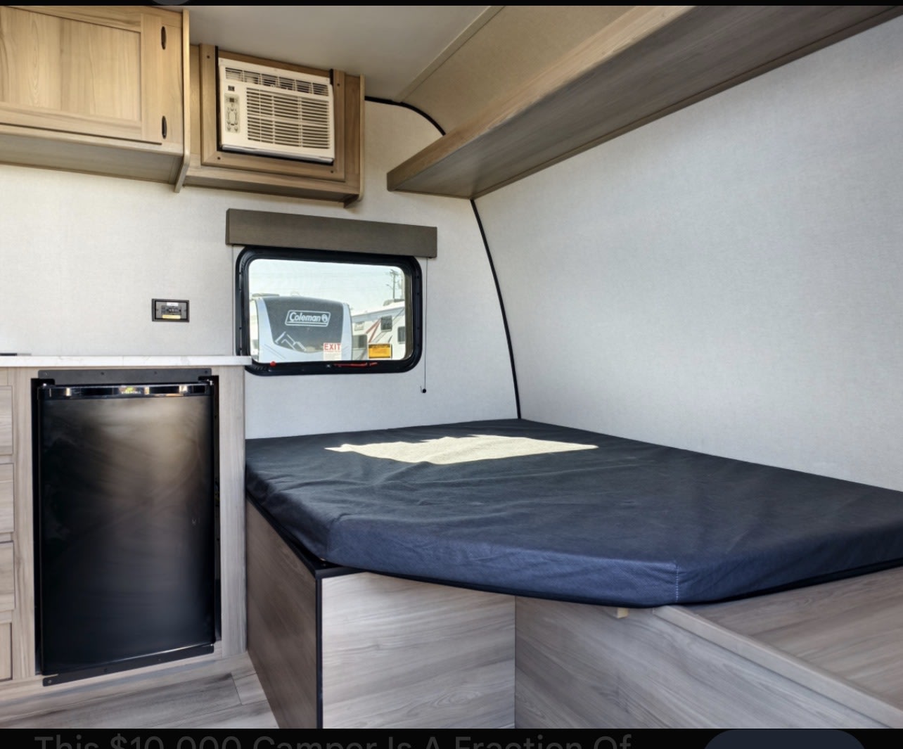 Compact rv rental interior featuring a comfortable bed, small refrigerator, AC unit, overhead cabinets, and a window.