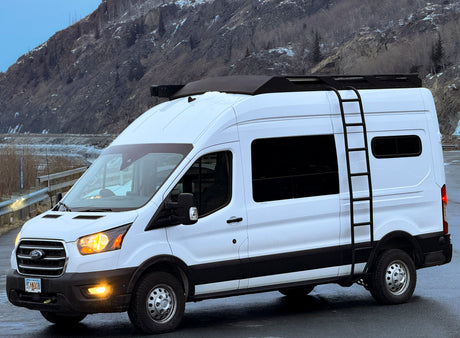 White Ford Transit rv rental camper van with roof rack and ladder, parked by snowy mountains.