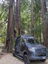 A dark grey Mercedes Sprinter rv rental parked on gravel in a towering redwood forest, offering a nature escape.