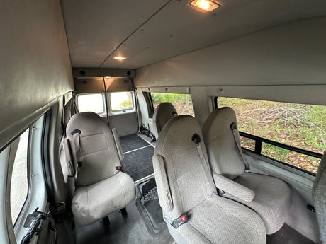RV rental interior: Rows of gray fabric passenger seats, overhead lights, and rear windows looking out to trees and bushes.