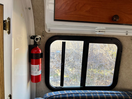 Essential RV rental safety: a fire extinguisher mounted beside a window with an outdoor view, cozy bed below.