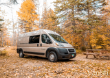 Autumn adventure: A modern tan RV rental, a converted camper van, is nestled in a colorful fall forest campsite with a picnic table.