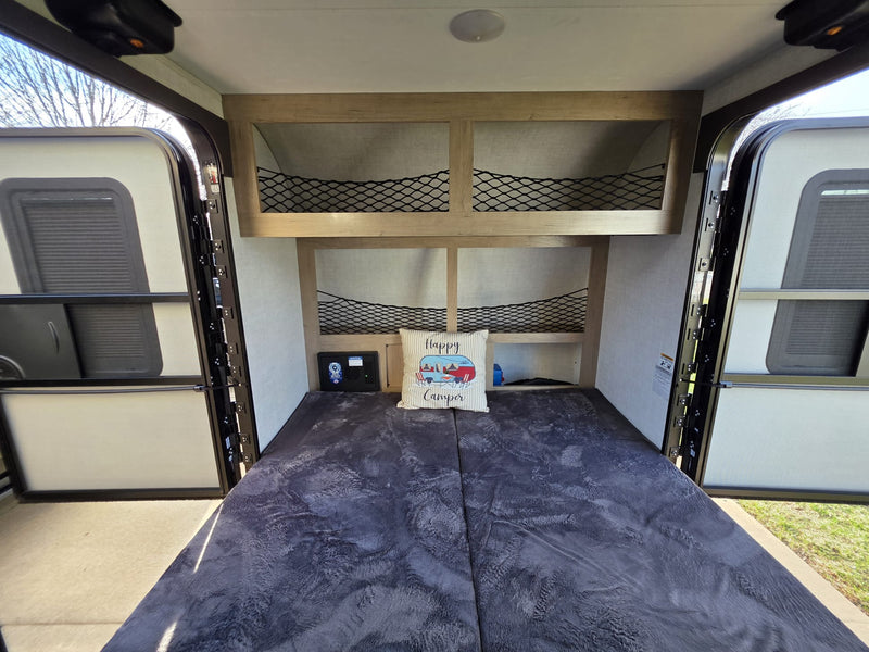 This RV rental shows a compact interior with a bed, "Happy Camper" pillow, overhead storage with netting, and open back doors. — photo 11