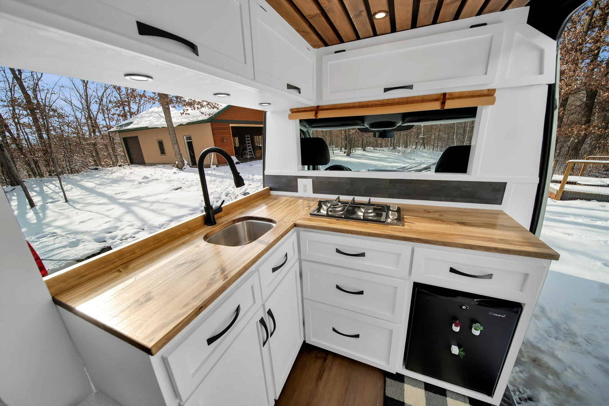 Sleek RV rental kitchen: wood counters, white cabinets, gas stove, sink, mini-fridge, with a snowy winter forest outside. — photo 9