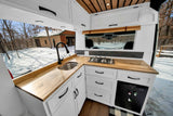 Sleek RV rental kitchen: wood counters, white cabinets, gas stove, sink, mini-fridge, with a snowy winter forest outside.