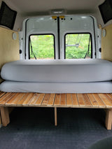 Inside an RV rental van: a cozy bed setup with wooden slats, stacked gray mattresses, and back windows.