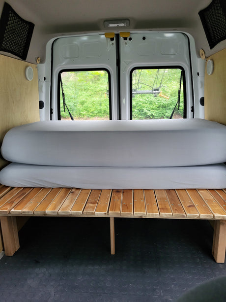Inside an RV rental van: a cozy bed setup with wooden slats, stacked gray mattresses, and back windows.