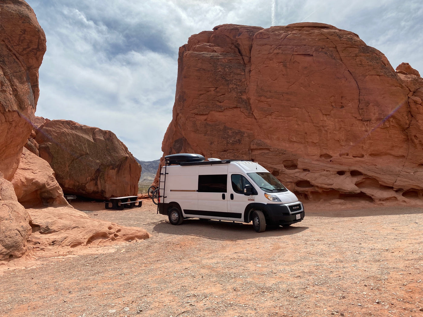 White RV rental camper van parked in a vibrant red rock desert, with large sandstone formations and a picnic table. Ideal for adventure.