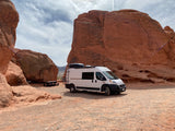 White RV rental camper van parked in a vibrant red rock desert, with large sandstone formations and a picnic table. Ideal for adventure.