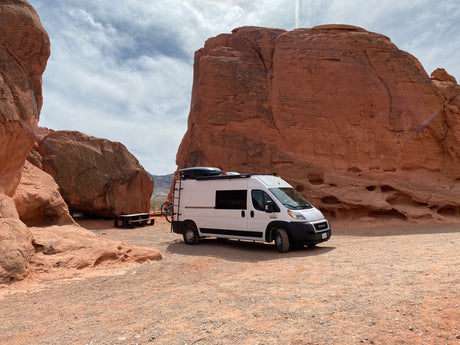 White RV rental camper van parked in a vibrant red rock desert, with large sandstone formations and a picnic table. Ideal for adventure.