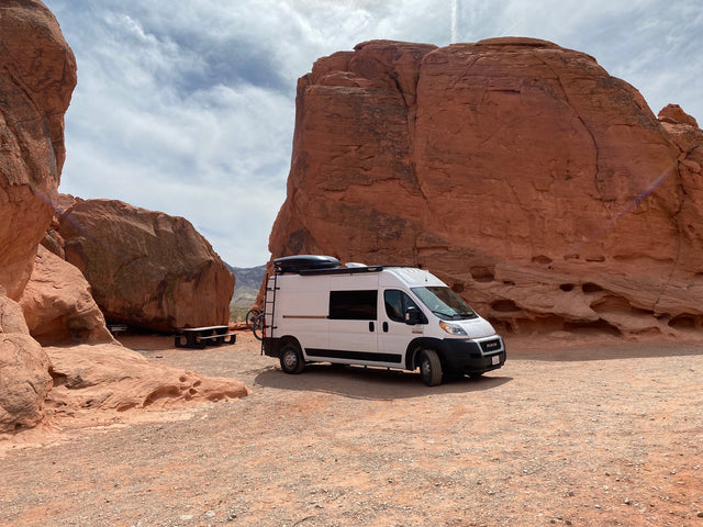 White RV rental camper van parked in a vibrant red rock desert, with large sandstone formations and a picnic table. Ideal for adventure.