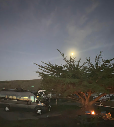 An RV rental camper van parked at night. A bright moon shines above a large tree, with a cozy campfire below. Perfect for camping.
