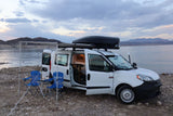 This RV rental camper van is parked by a scenic lake at dusk, with an awning, chairs, and table set up for camping.