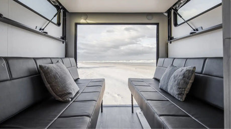 RV rental: Plush black seating inside, offering a panoramic view of a serene sandy beach and ocean under a cloudy sky.