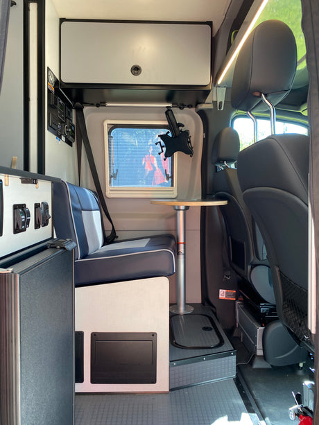 Compact RV rental interior with seating, a small table, overhead storage, and control panel. Well-lit and organized.