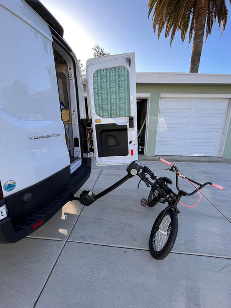 White RV rental van, open doors revealing interior, holds a BMX bike on its rear rack. Ready for adventure on a sunny day.