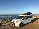 White Ford Transit Connect RV rental with a roof box parked seaside, ideal for beach trips and family adventures.