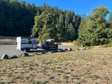 RV rental: A white and blue travel trailer towed by an SUV, parked on asphalt beside a forest on a sunny day.