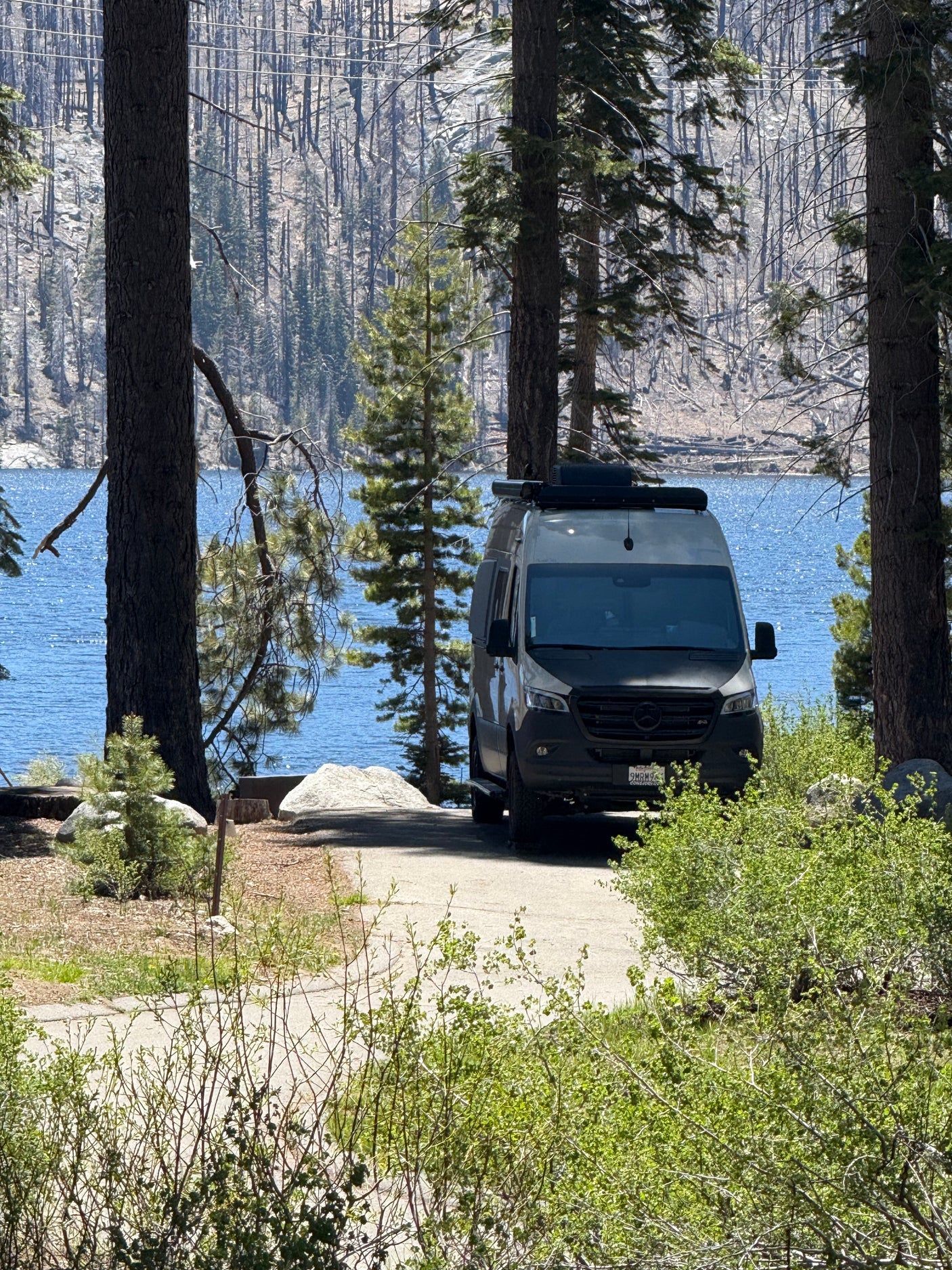 RV rental camper van by a sparkling blue lake, framed by pines, with distant mountains and a burnt forest.