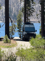 RV rental camper van by a sparkling blue lake, framed by pines, with distant mountains and a burnt forest.
