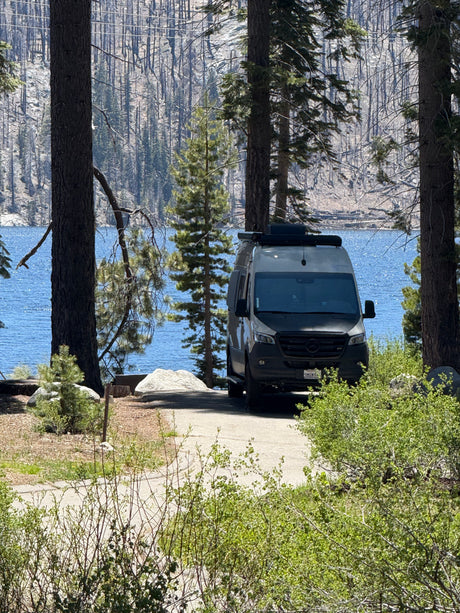 RV rental camper van by a sparkling blue lake, framed by pines, with distant mountains and a burnt forest.