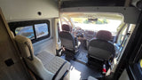 RV rental camper van interior. View from rear showing driver, passenger, and a side-facing rear seat, with a side window.