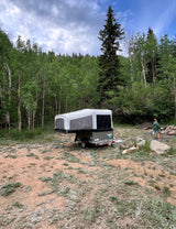 Pop-up camper RV rental set up in a wooded campsite with a child walking near a fire pit, surrounded by trees.