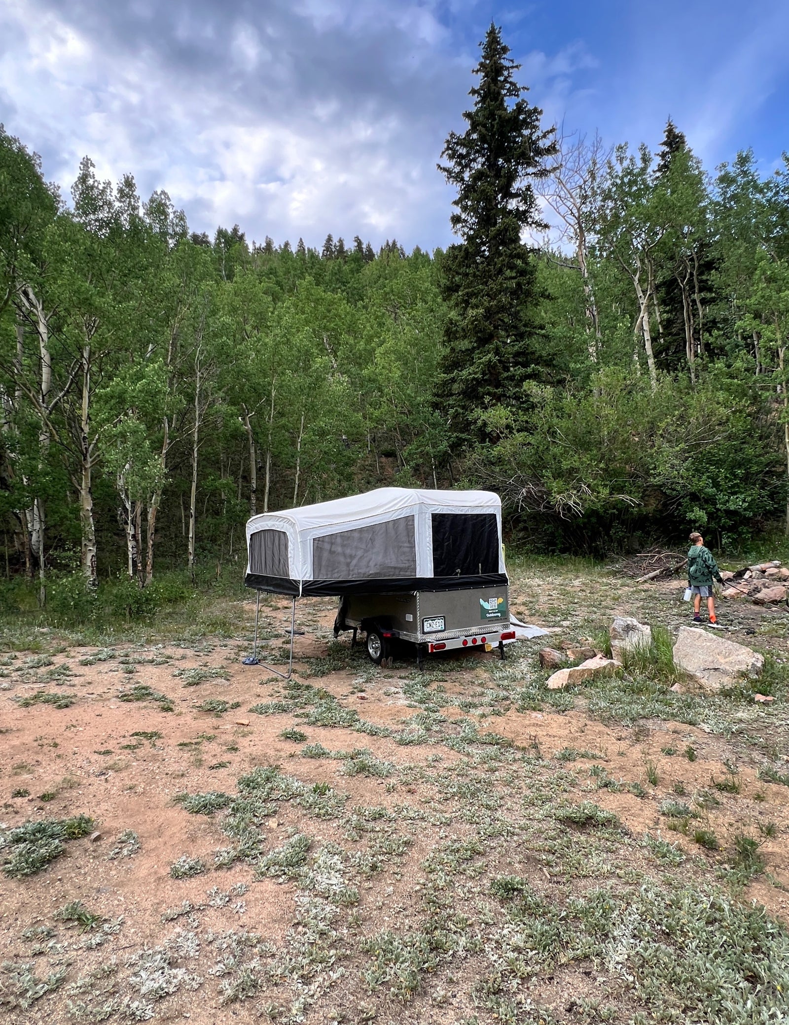 Pop-up camper RV rental set up in a wooded campsite with a child walking near a fire pit, surrounded by trees.
