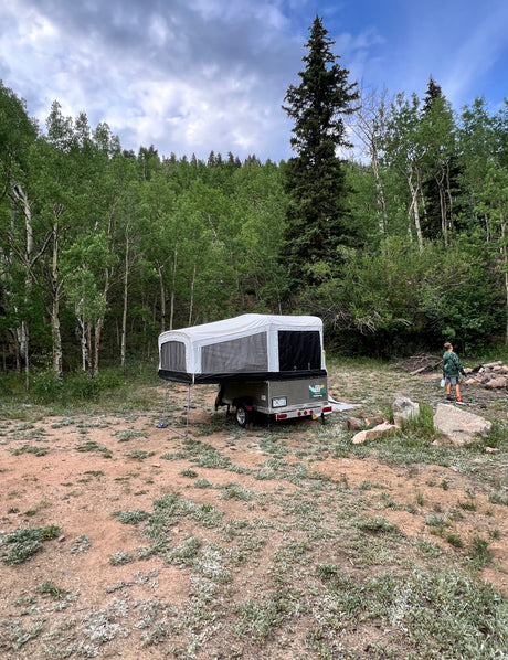 Pop-up camper RV rental set up in a wooded campsite with a child walking near a fire pit, surrounded by trees.