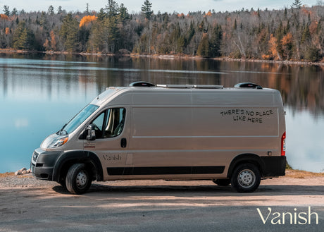Light beige RV rental parked lakeside, autumn trees reflecting. The van reads: 'THERE'S NO PLACE LIKE HERE'.
